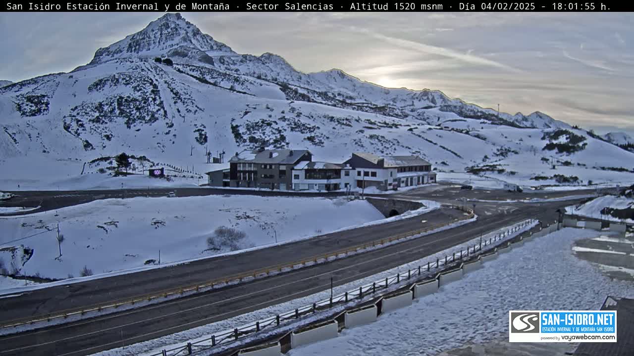 A snow-covered mountain slope with a road and buildings below, under a twilight sky.