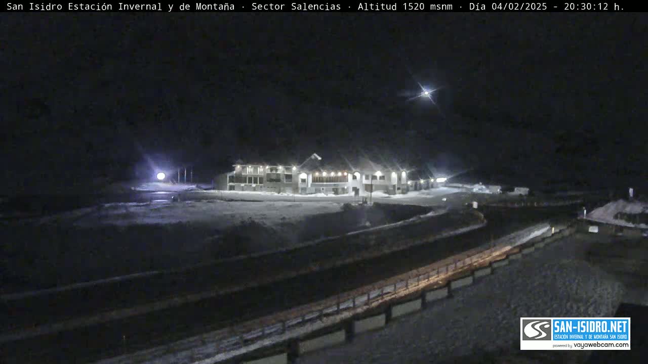 A snow-covered, nighttime scene shows a large building illuminated with lights, next to a road, under a clear, dark sky with a bright star visible.
