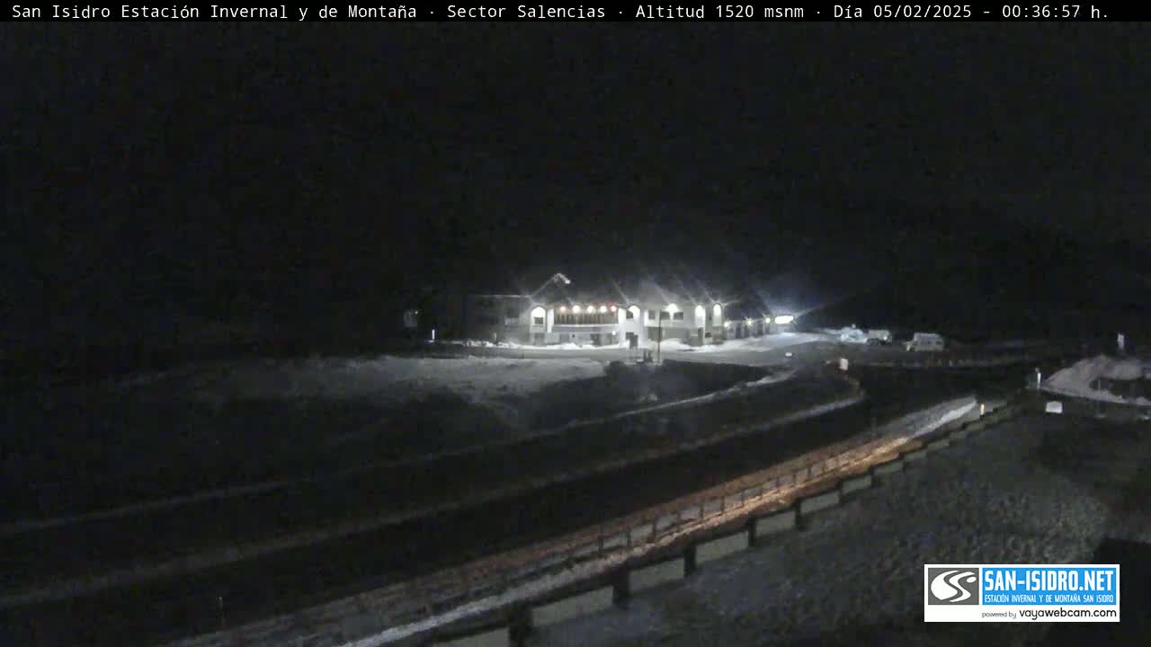 A snow-covered landscape at night, featuring a brightly lit building and a road, under dark, possibly snowy conditions.