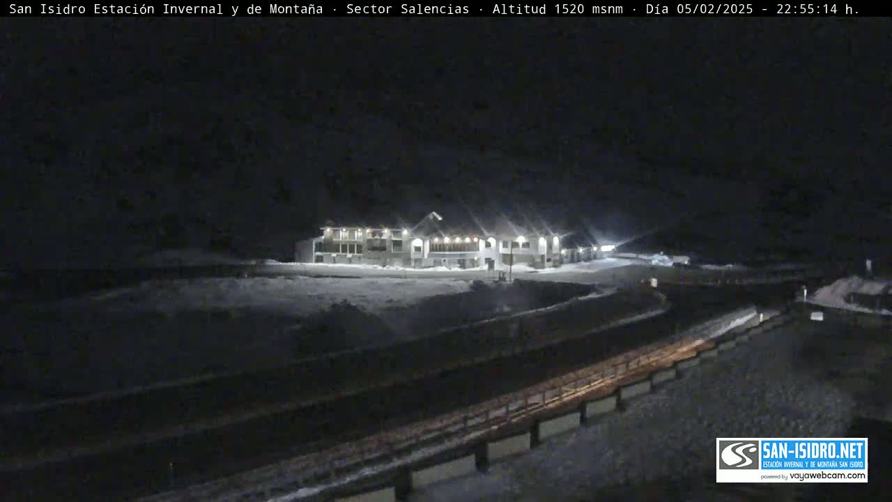 A snow-covered landscape at night, illuminated by the lights of a building in the distance.