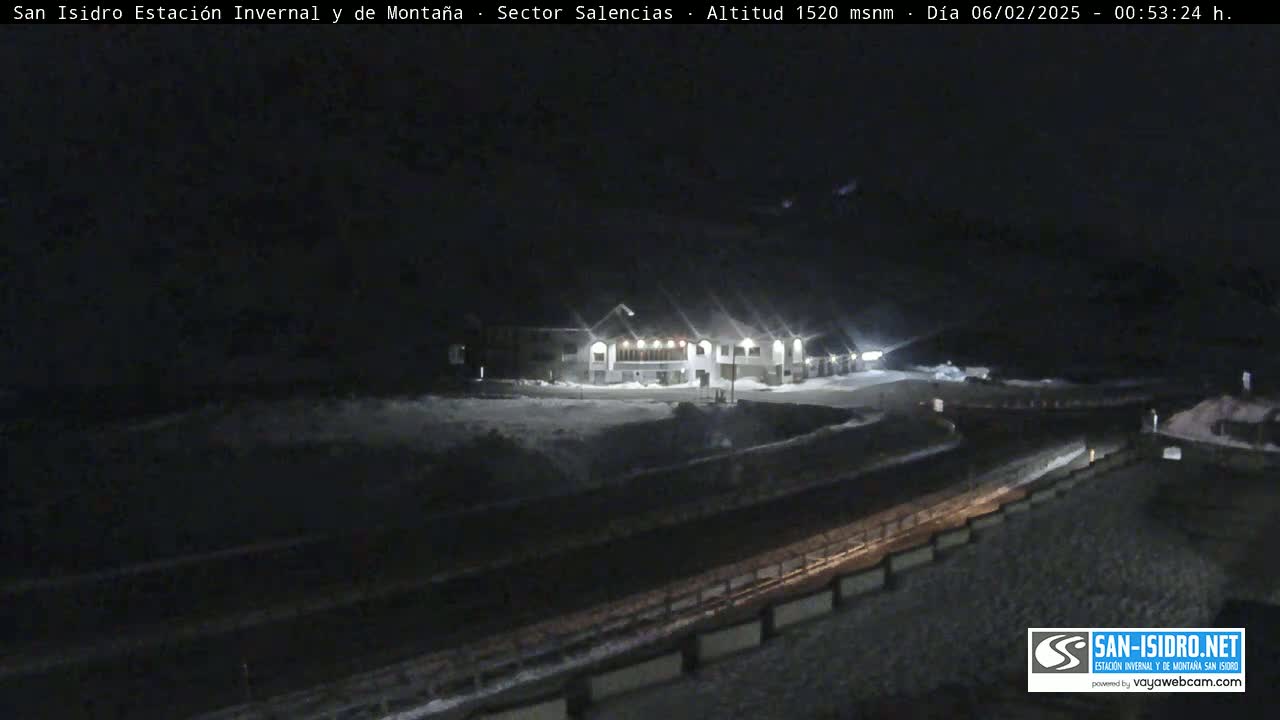 A snow-covered, nighttime scene shows a building lit up with lights, a road, and what appears to be a retaining wall.