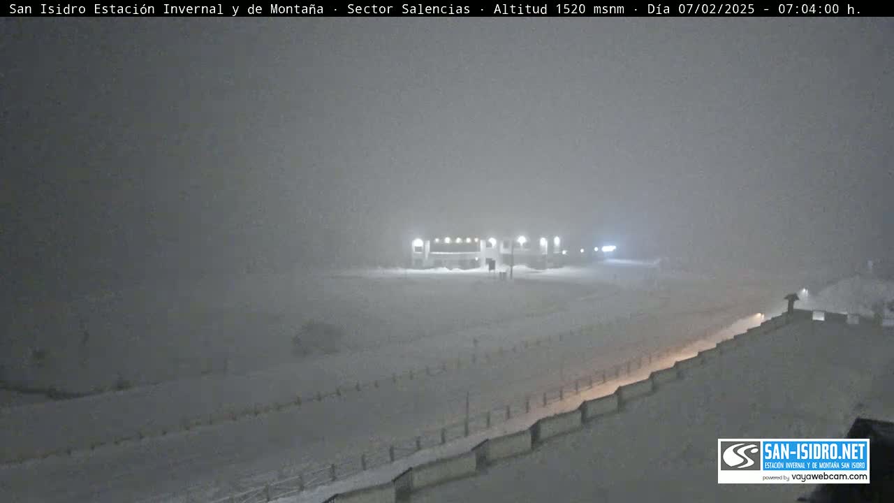 A snow-covered landscape at night, featuring a building with lights and a road partially obscured by heavy snowfall.
