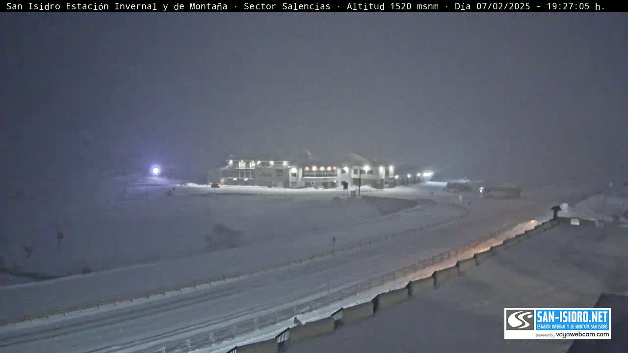 A snow-covered landscape at night shows a building lit up in the distance under heavy snowfall.