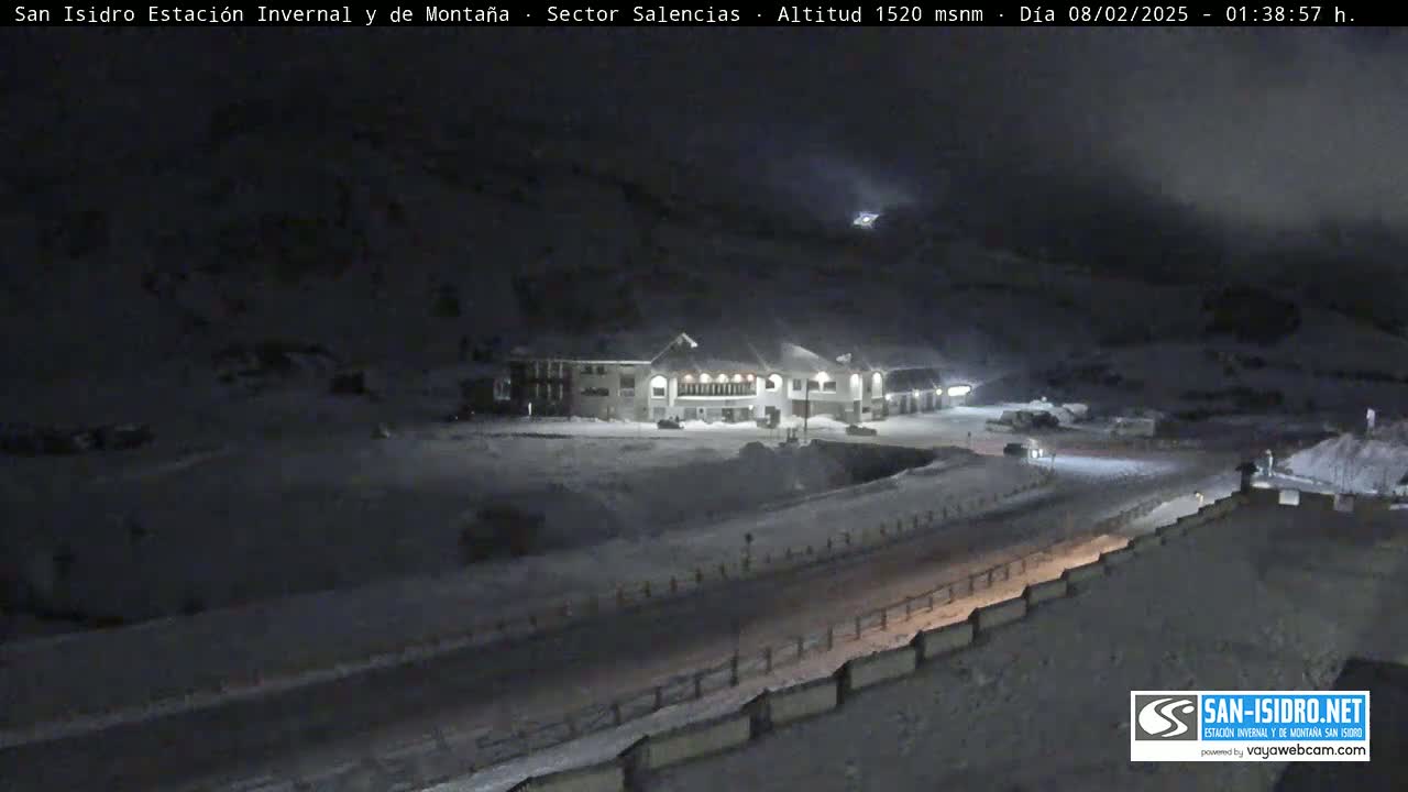 A snow-covered landscape at night, featuring a lit building and a road, under a dark, cloudy sky.