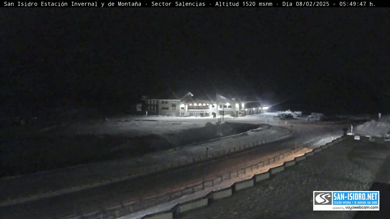 A snow-covered landscape at night, featuring a lit building and a road, is illuminated by artificial light.