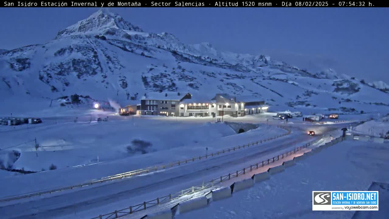 A snow-covered mountainside with a building complex and a road in the foreground under a twilight sky.