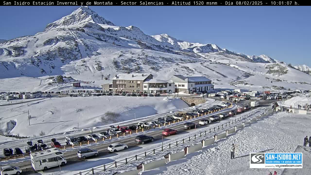 A snow-covered mountain landscape with a building and numerous cars parked in a large lot, under a clear, sunny sky.
