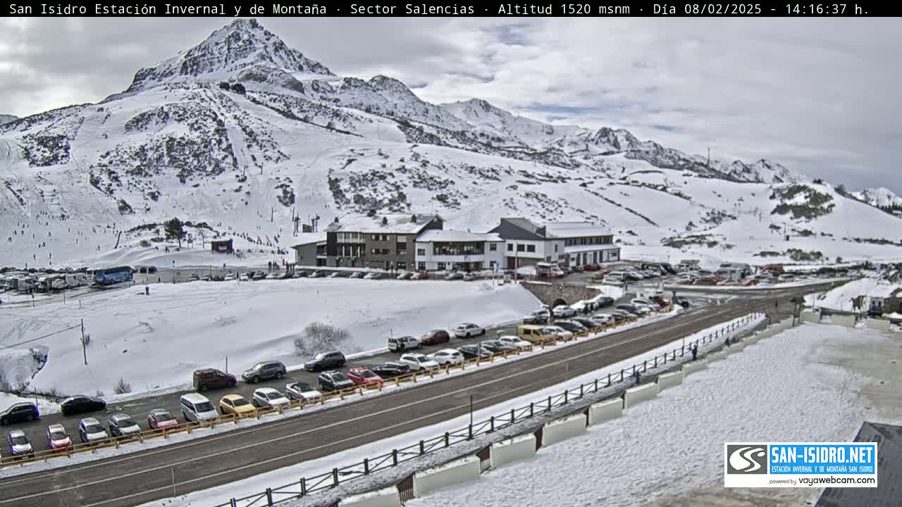 A snow-covered mountain slope with ski runs, a building complex, and a parking lot full of cars, all under a cloudy sky.