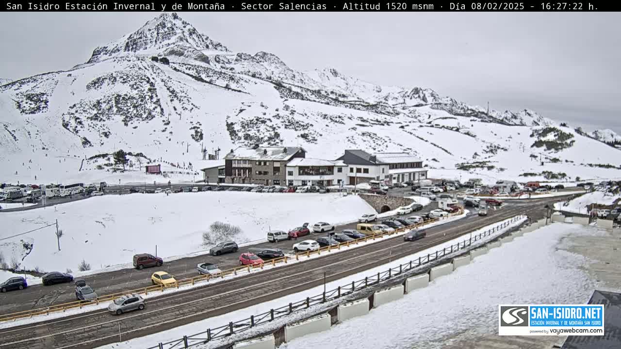 A snow-covered mountainside with ski slopes, a parking lot full of cars, and a building complex is visible under an overcast sky.