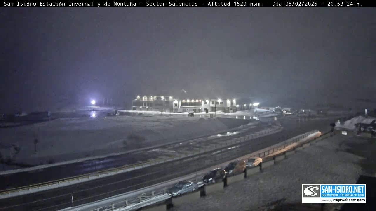 A snow-covered landscape at night features a large building near a road with several cars parked alongside it, under heavy snowfall.