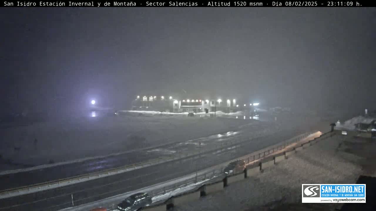 A nighttime, snowy scene shows a highway and a building with lights, under heavy fog or snow.