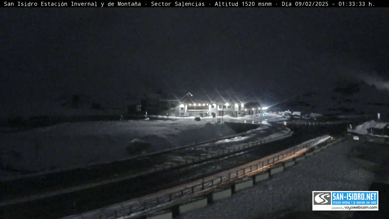 A snow-covered nighttime scene shows a building complex illuminated by lights, with a road curving through the area.