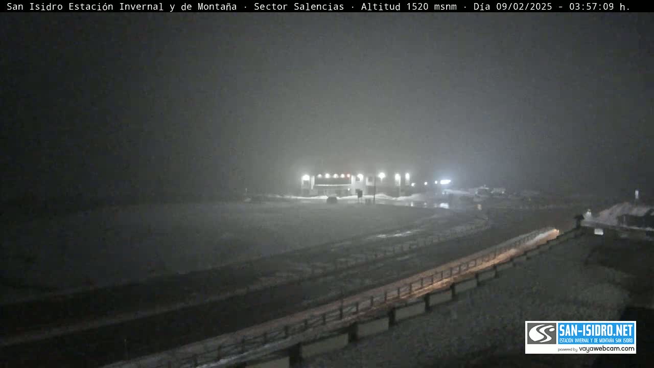 A nighttime, snowy scene shows a building complex near a road, under foggy conditions.