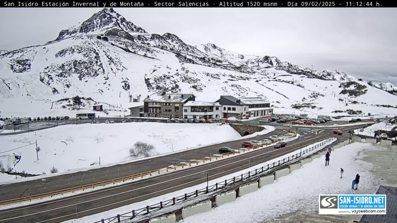 A snow-covered mountain slope with a ski resort building and parking lot, a road, and several cars, under an overcast sky.