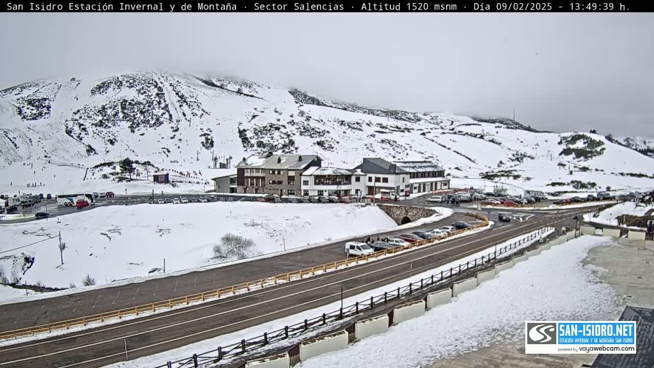 A snow-covered mountainside with ski slopes, a cluster of buildings, and a road with parked cars, under an overcast sky.