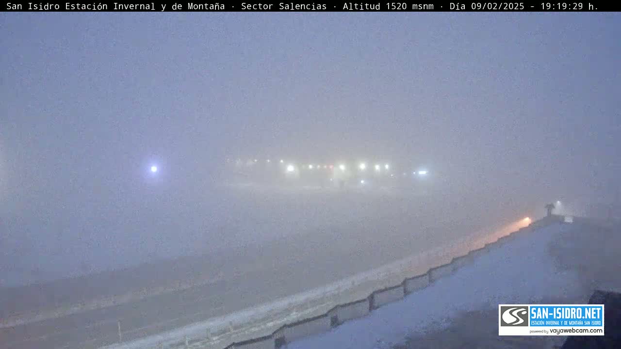 A snow-covered landscape is shrouded in heavy fog at night, with dimly lit structures barely visible in the distance.