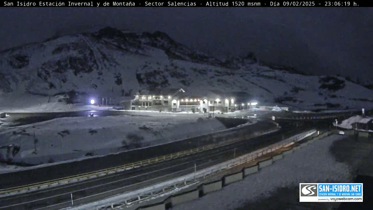 A snow-covered mountainside at night, illuminated by lights from a building near a road.
