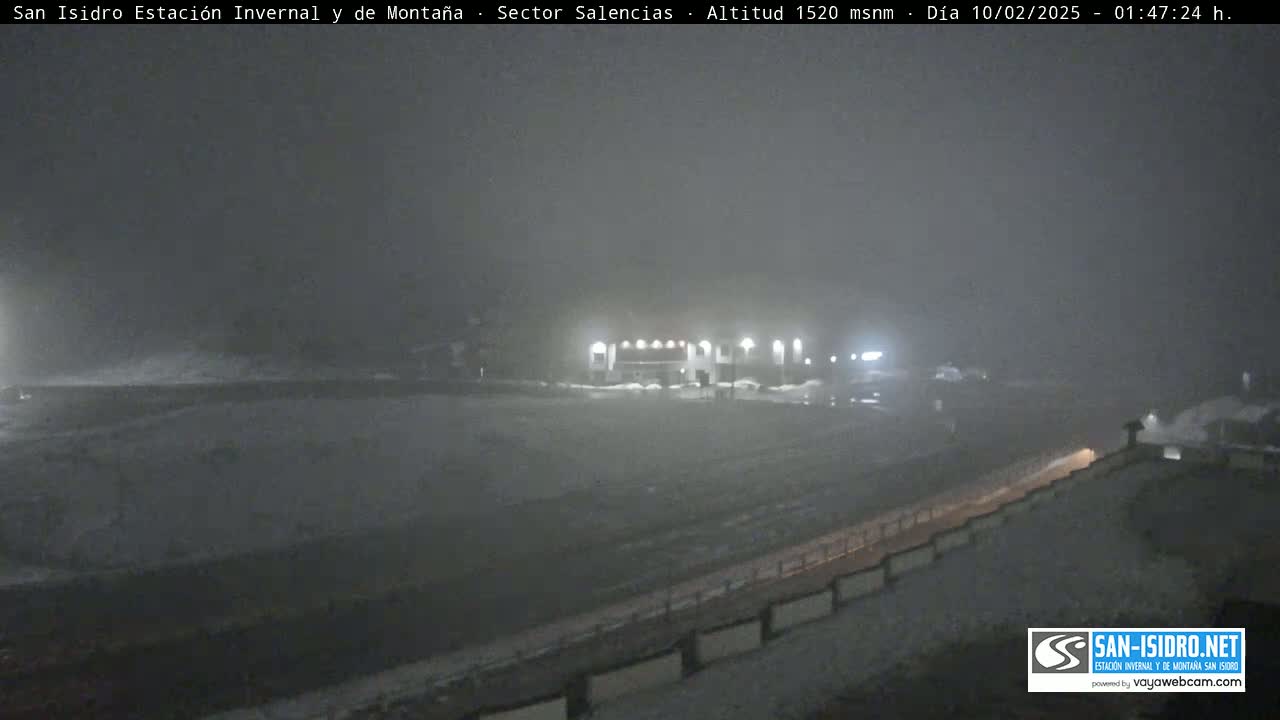 A snow-covered landscape at night, showing a building with lights and a road partially obscured by blowing snow.