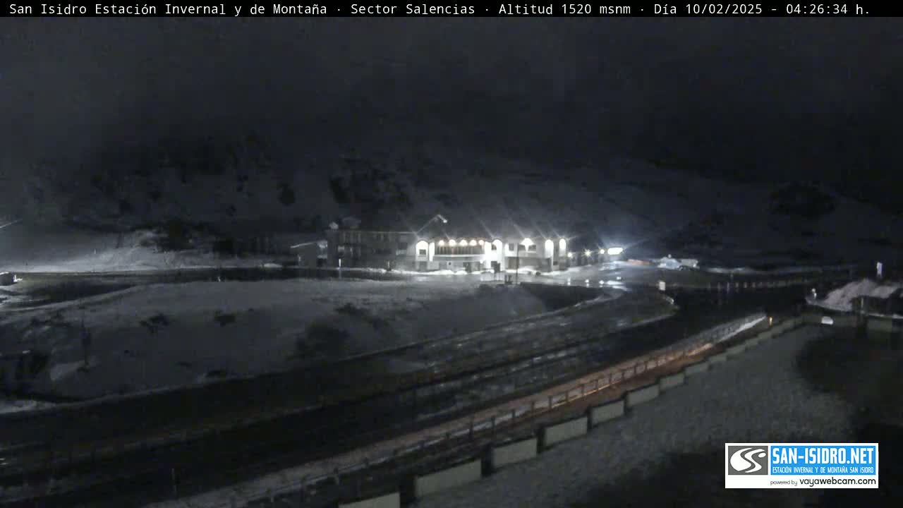 A snow-covered mountain landscape at night, illuminated by the lights of a building near a roadway.