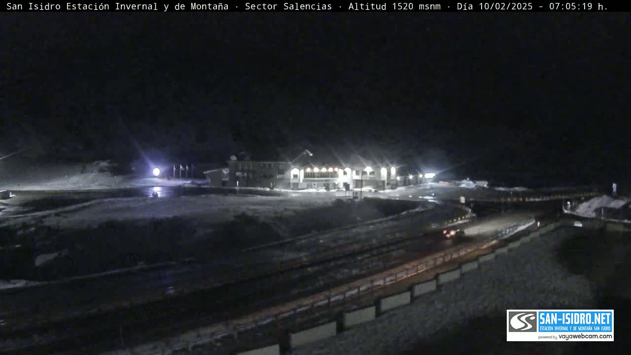 A nighttime view of a snow-covered mountain area showing a building with lights on and a road with a vehicle, under dark conditions.