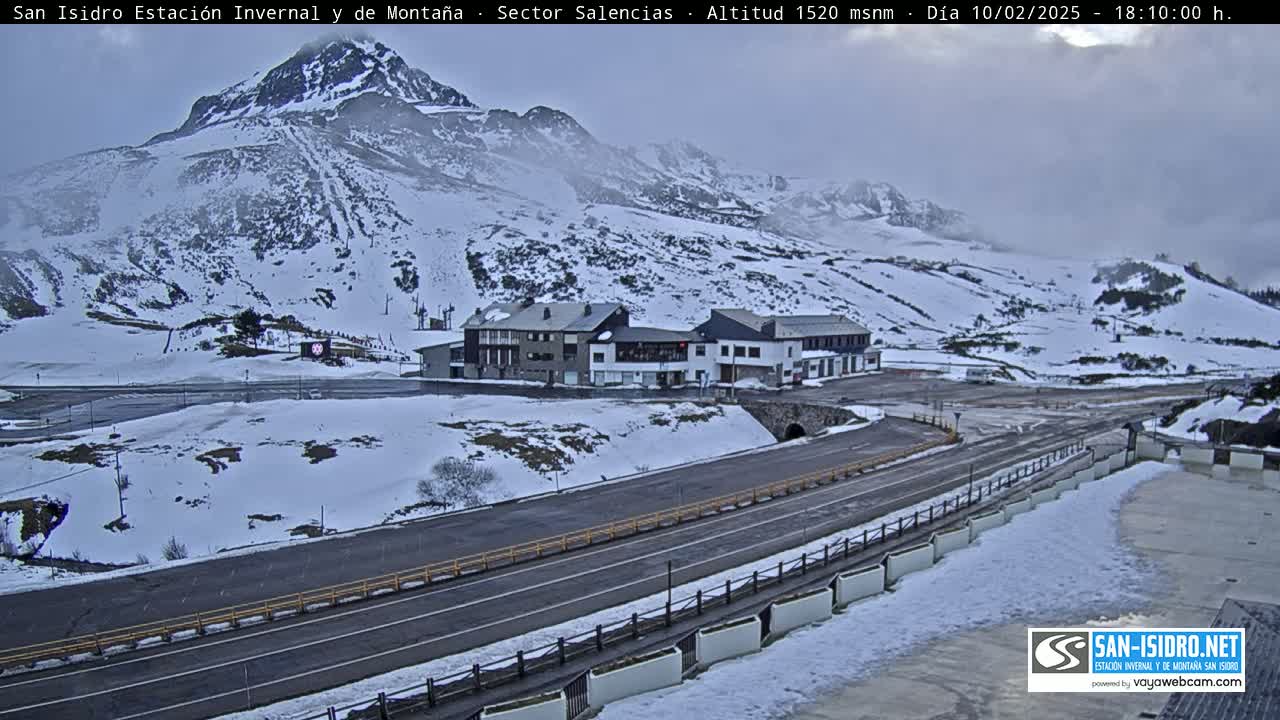 A snow-covered mountain landscape with a road and several buildings near the base, under an overcast sky.