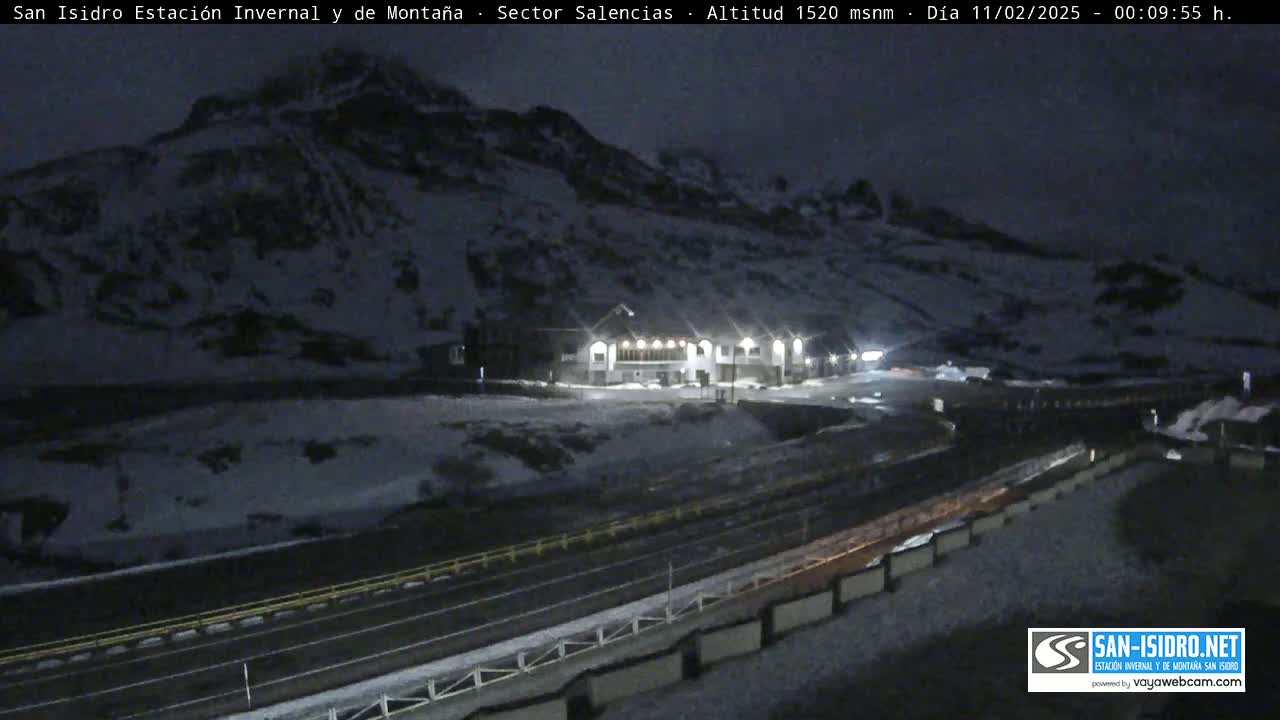 A snow-covered mountainside at night is illuminated by the lights of a building near a road.