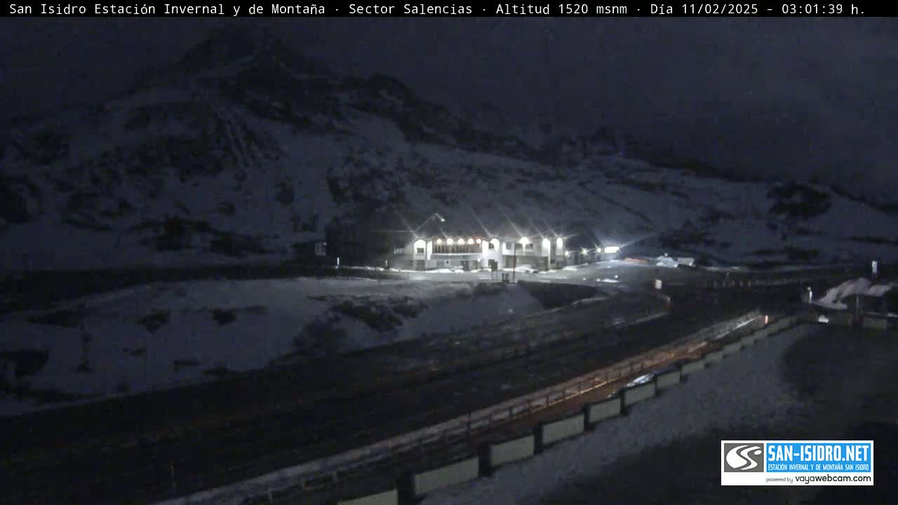 A snow-covered mountainside at night is illuminated by the lights of a building below, with a road and barrier visible in the foreground.