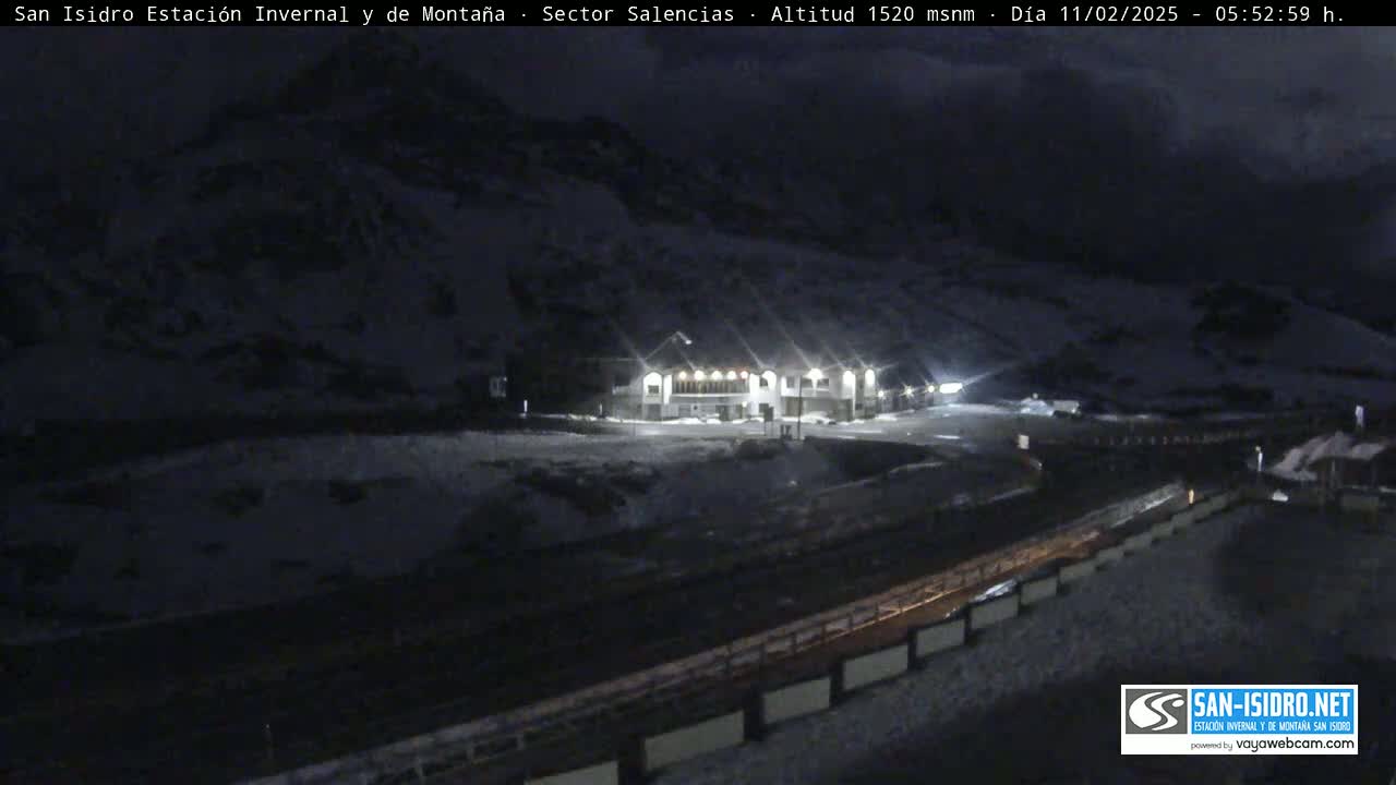 A nighttime, snow-covered mountain scene shows a lit building near a road, with a dark, snow-covered mountainside behind it.