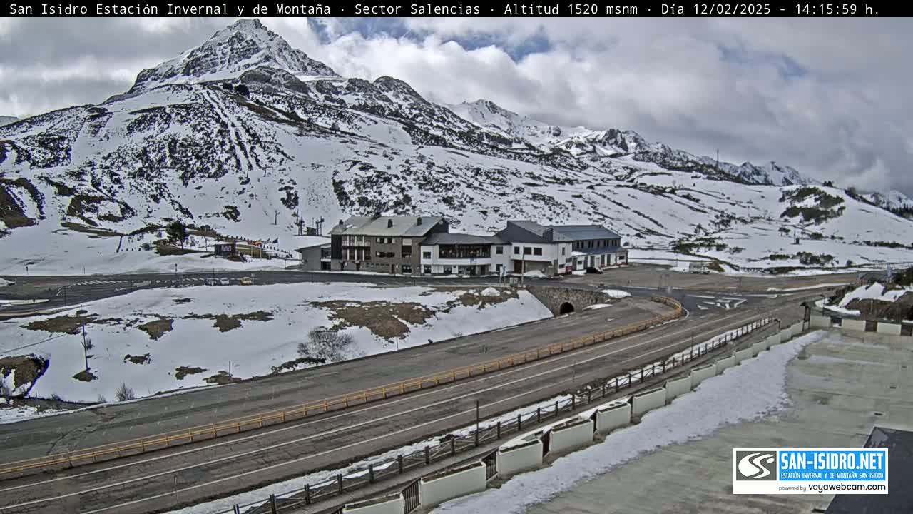 A snow-covered mountainside with several buildings near a roadway under a partly cloudy sky.