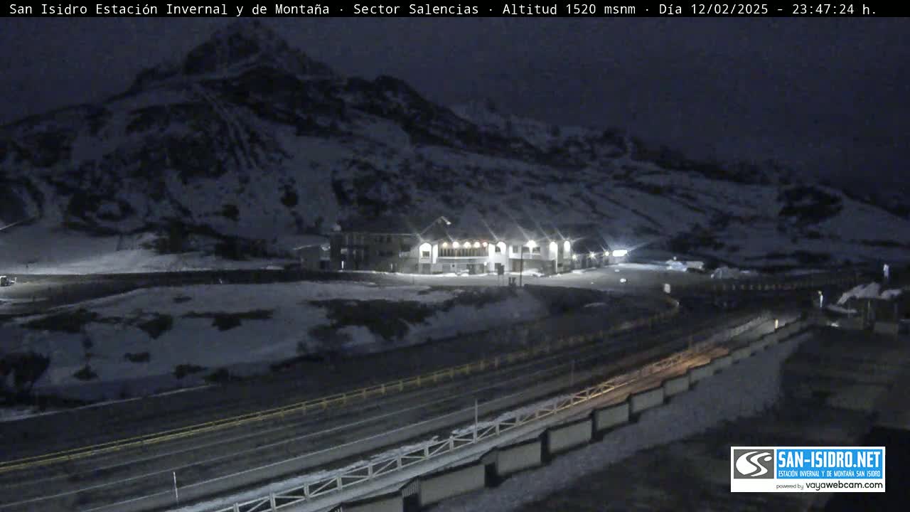 A nighttime, snow-covered mountain scene shows a building with lights on near a road.
