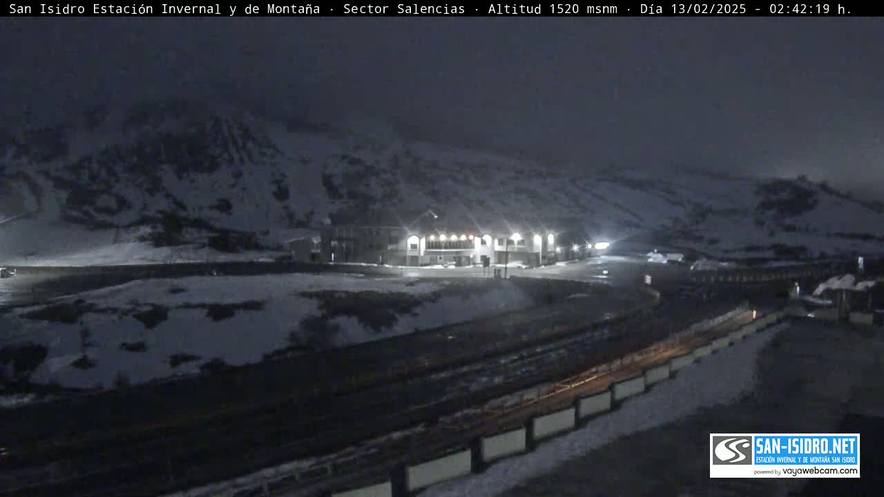 A snow-covered mountainside at night is illuminated by lights from a building and vehicles near a road.