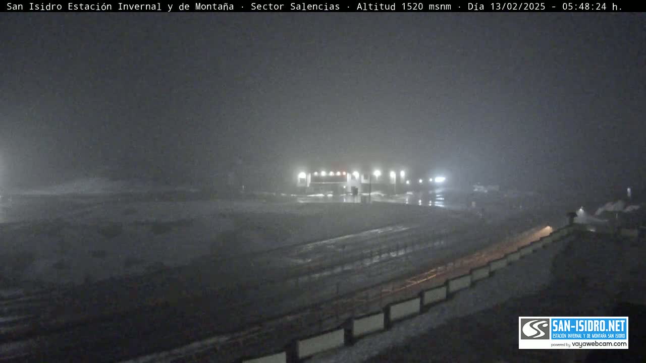 A nighttime, foggy scene shows a snow-covered road leading to a building complex with lights, under hazy conditions.