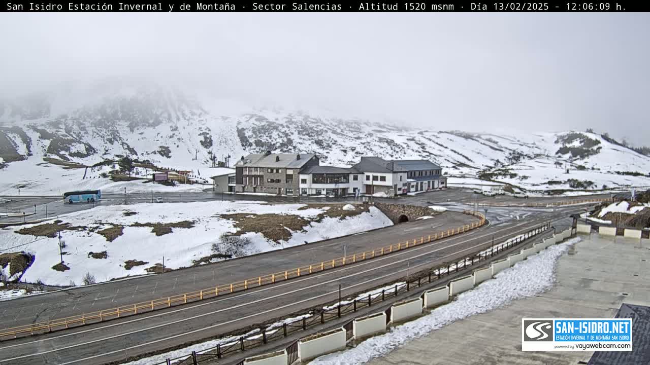 A snow-covered mountain landscape with a road, buildings, and a bus in a parking lot under overcast skies.