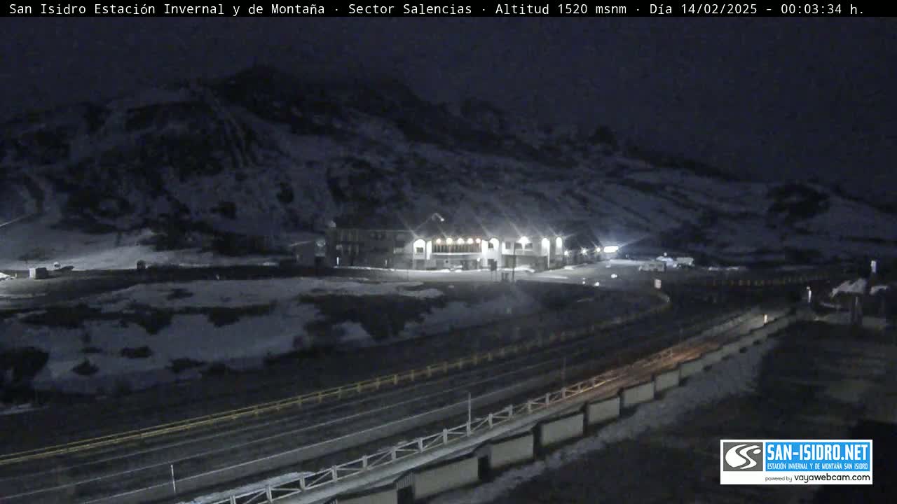 A nighttime view of a snow-covered mountainside with a building illuminated by lights and a road below.