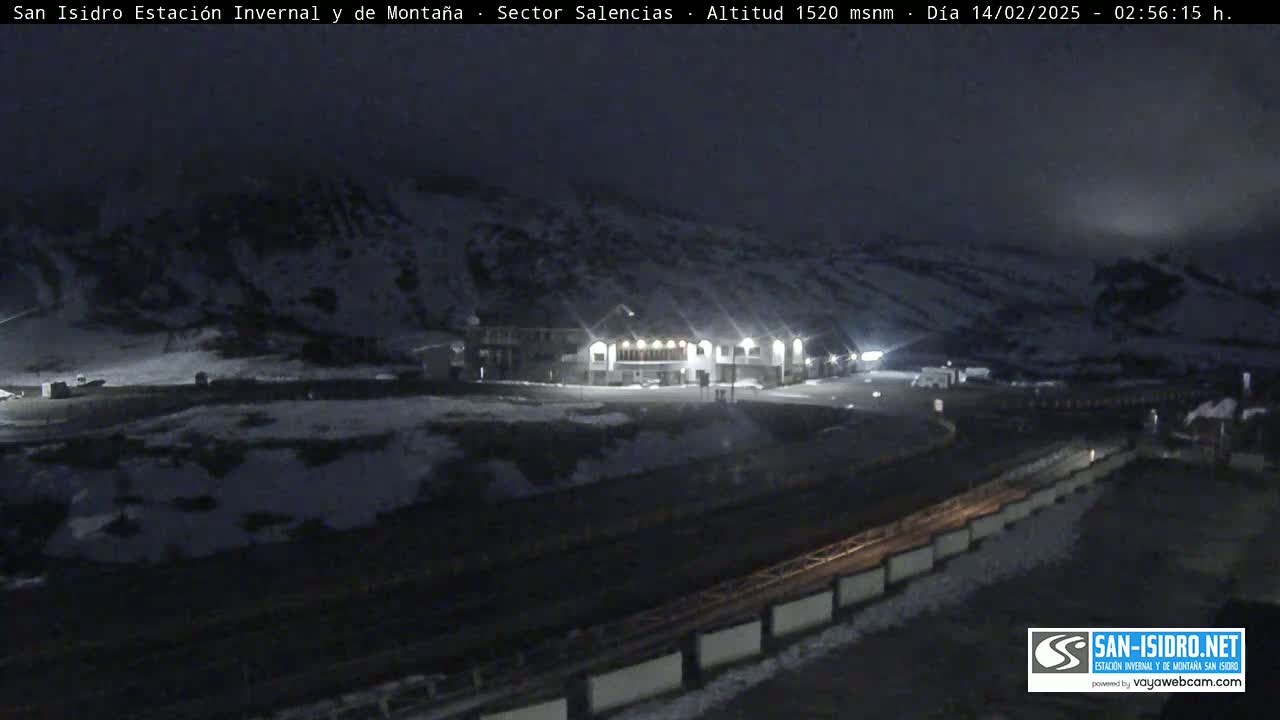 A nighttime, snow-covered mountain landscape with a lit building and parking lot, under a dark, cloudy sky.