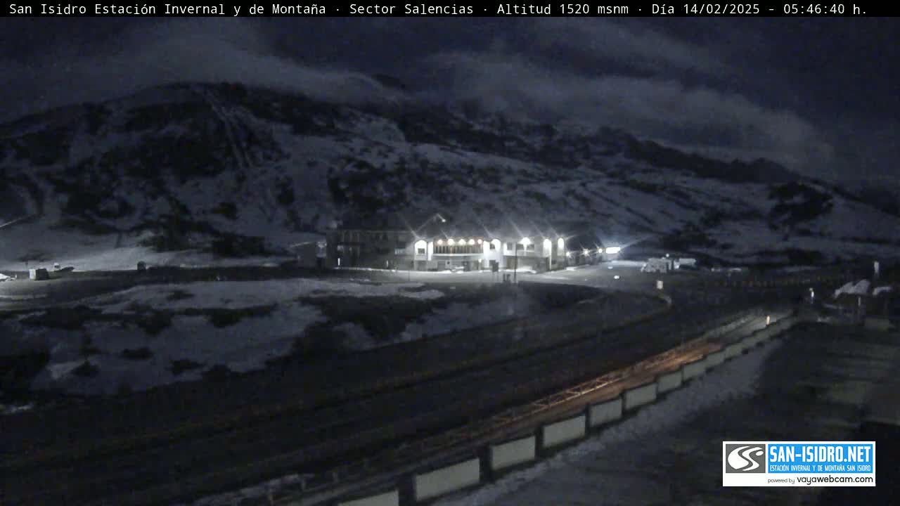 A snow-covered mountainside at night is visible, with a brightly lit building near a roadway below under a dark, cloudy sky.