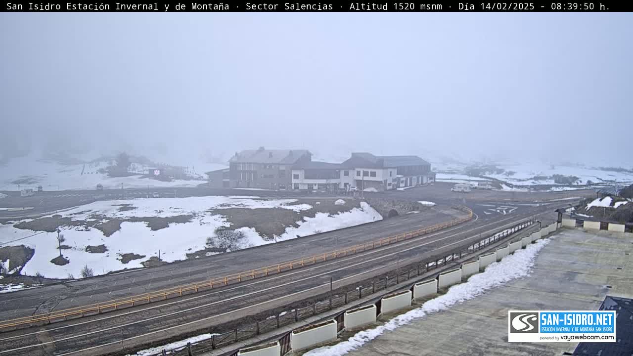 A foggy, snowy mountaintop scene shows a road curving past a cluster of buildings, with patches of snow covering the ground.