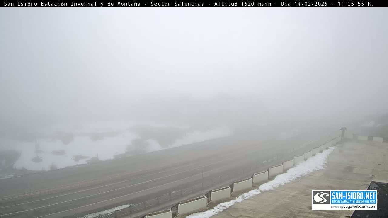 A foggy, snowy scene shows a road and a low wall with patches of snow on top.