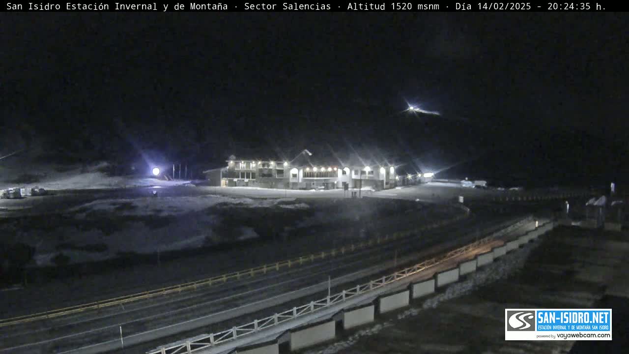 A nighttime, snow-covered scene shows a long building with lights on, a road, and a guardrail, under a dark sky.