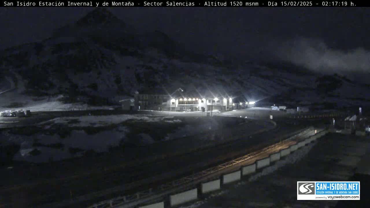 A nighttime view of a snow-covered mountainside with a lit building and a road below, under a cloudy sky.