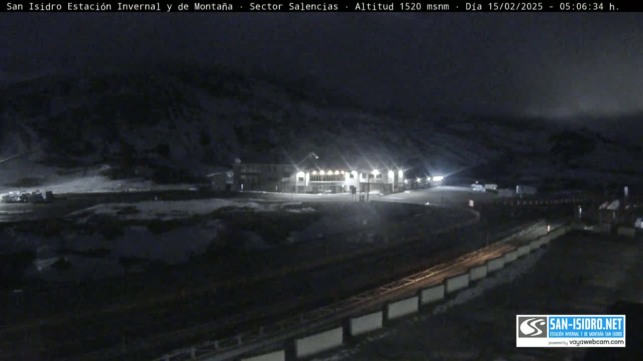 A nighttime, snowy scene shows a lit-up building in a mountain valley under a dark, overcast sky.