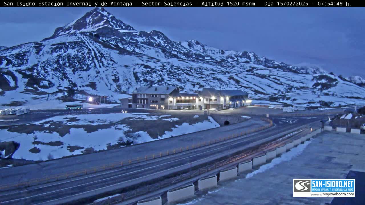 A snow-covered mountainside at twilight, with a roadway curving past a building complex and parking area.
