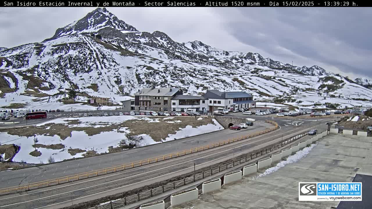 A mountainside ski resort with patchy snow cover under an overcast sky, showing a parking lot and buildings near a road.