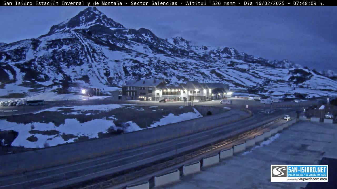 A snow-covered mountainside with a building complex and a roadway at its base under a twilight sky.