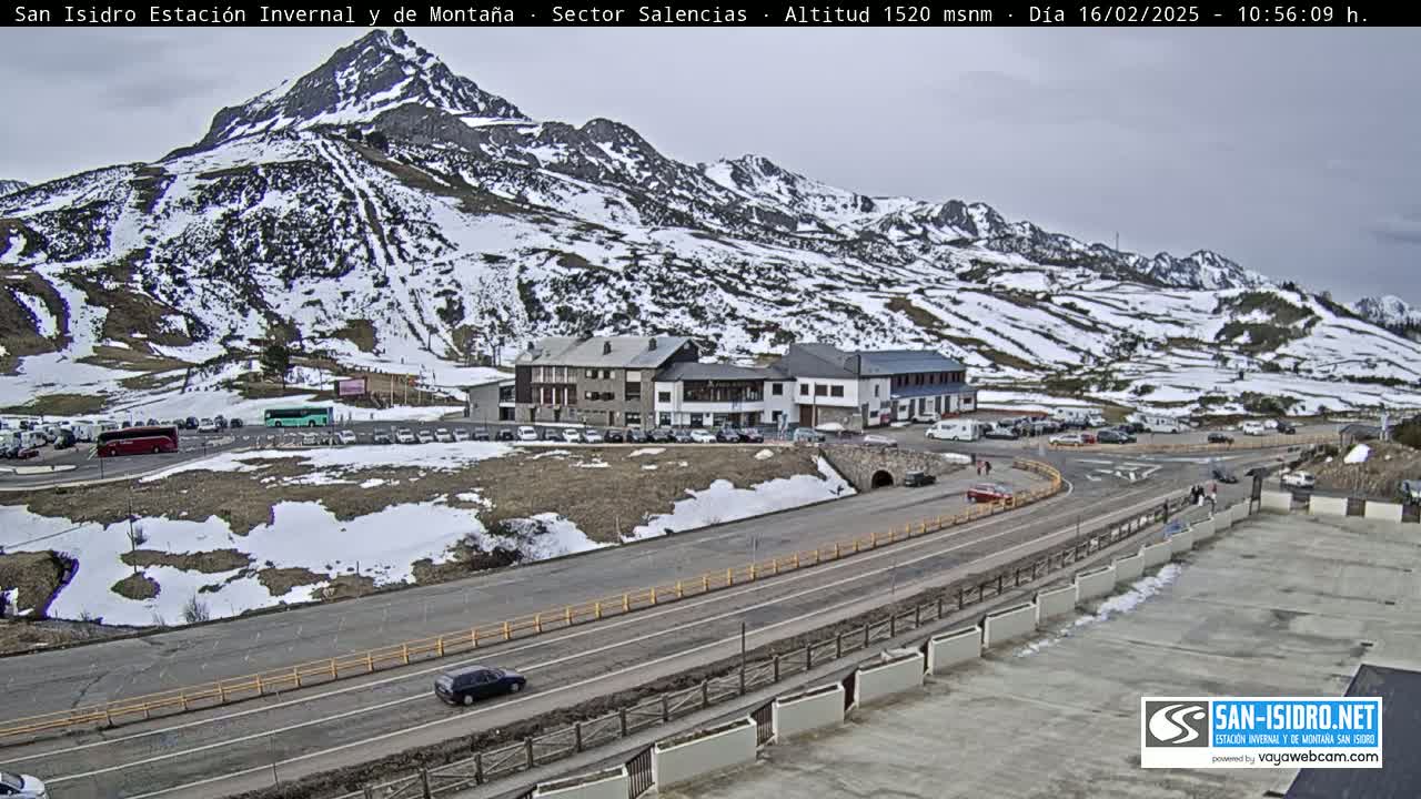 A mountain road winds through a snow-dusted mountain landscape, past a parking lot and buildings, under an overcast sky.