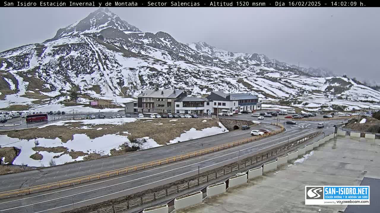 A snow-dusted mountainside overlooks a roadway with several vehicles parked near a cluster of buildings, under an overcast sky.