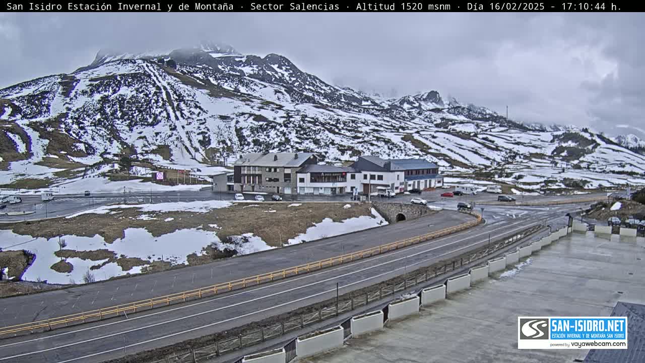 A snow-dusted mountainside overlooks a roadway and parking lot near several buildings, under an overcast sky.