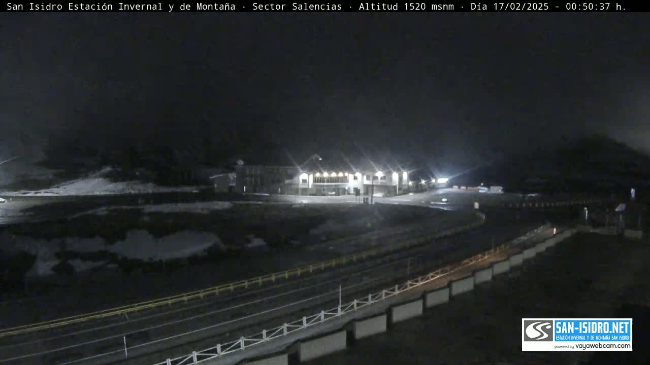 A nighttime view of a snow-covered mountain area showing a building complex illuminated with lights, surrounded by a dark road and a low fence.
