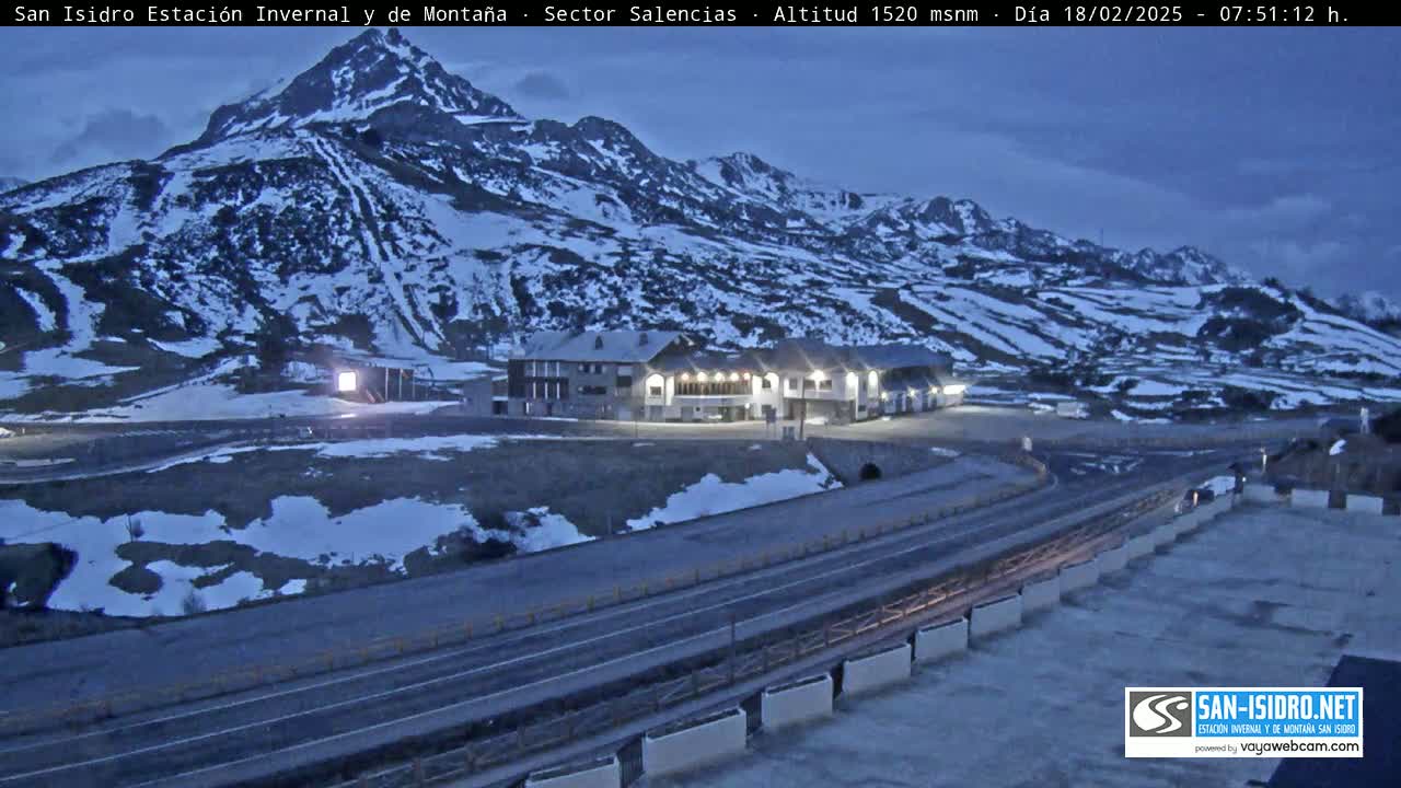 A snow-covered mountainside at dusk overlooks a road and a building complex near a mountain pass.