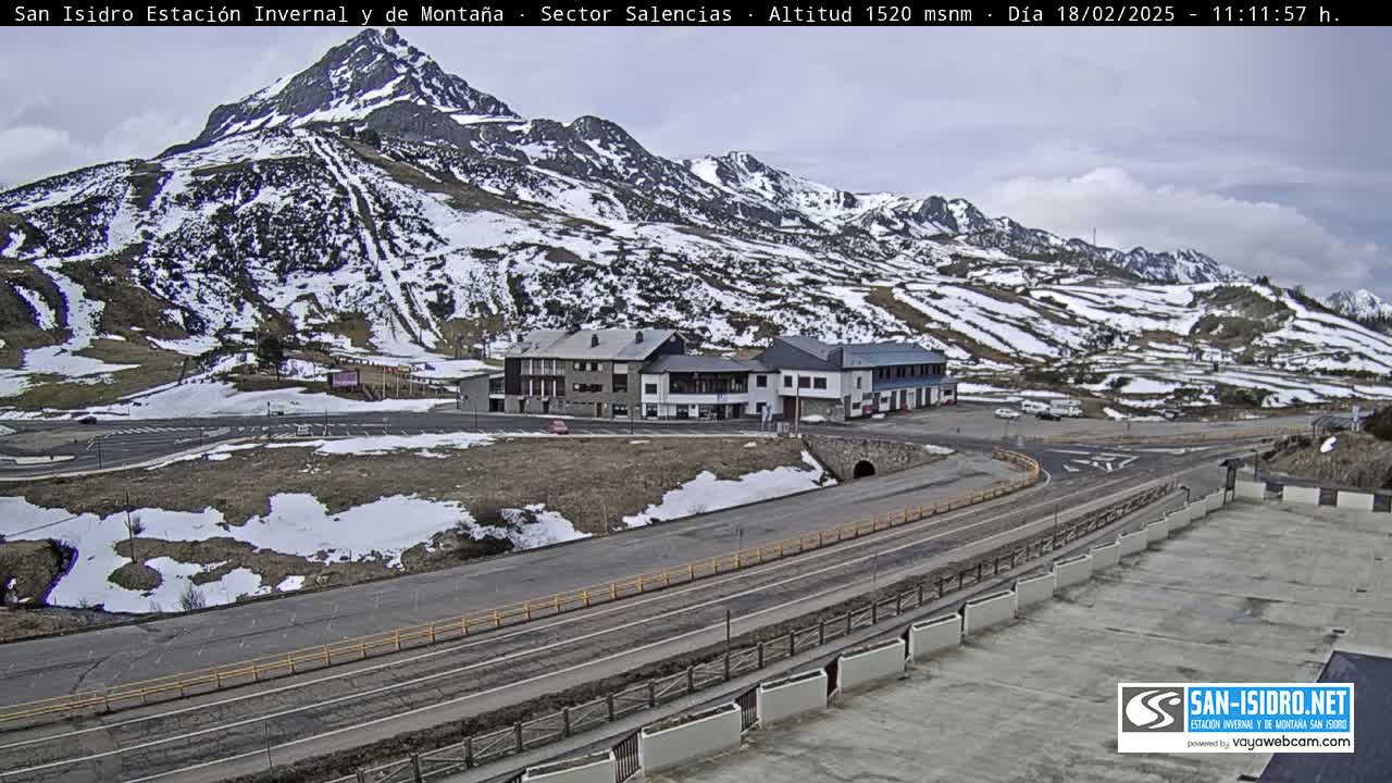A mountainside ski resort with patchy snow cover is visible under an overcast sky, with a road and several buildings situated in the foreground.
